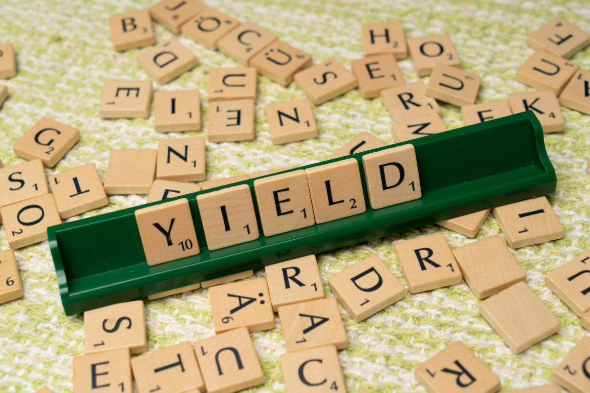 Fichas de Scrabble formando la palabra 'YIELD' en un soporte verde con letras dispersas en el fondo